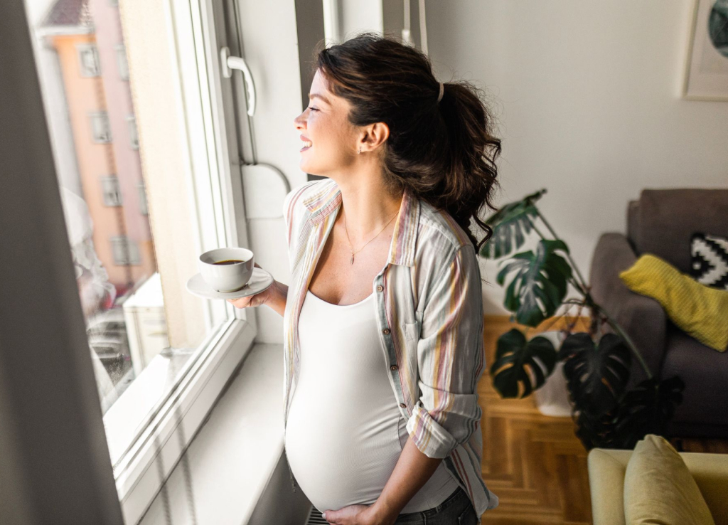 A pregnant woman happily looks out the window while holding a cup of coffee.